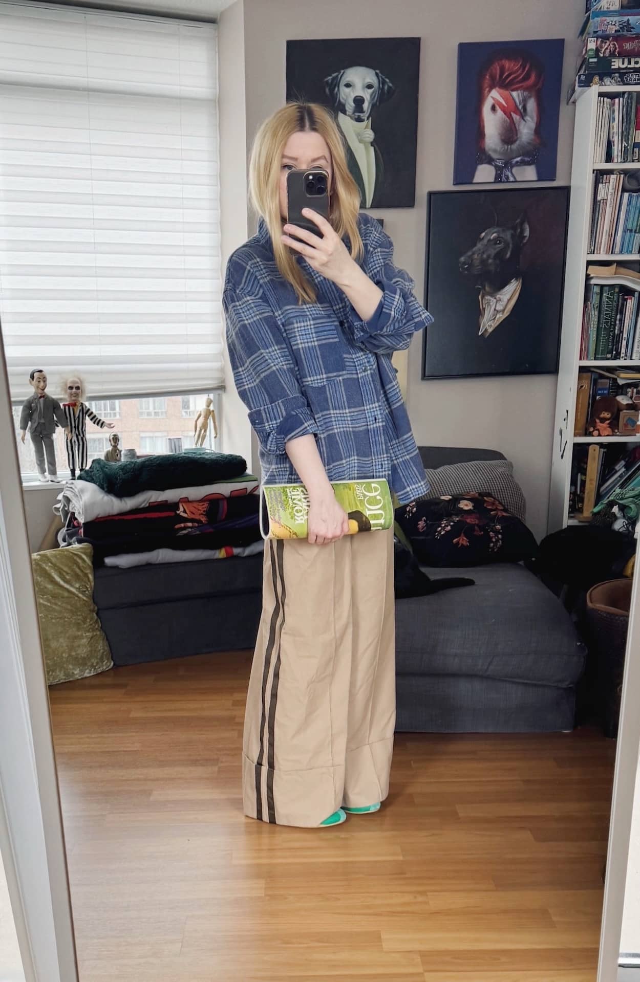 Sara of livelovesara is standing in front of a mirror taking a photo of her outfit. She is wearing a blue plaid shirt with wide leg tan trousers, a vintage magazine clutch, and green Adidas sneakers.