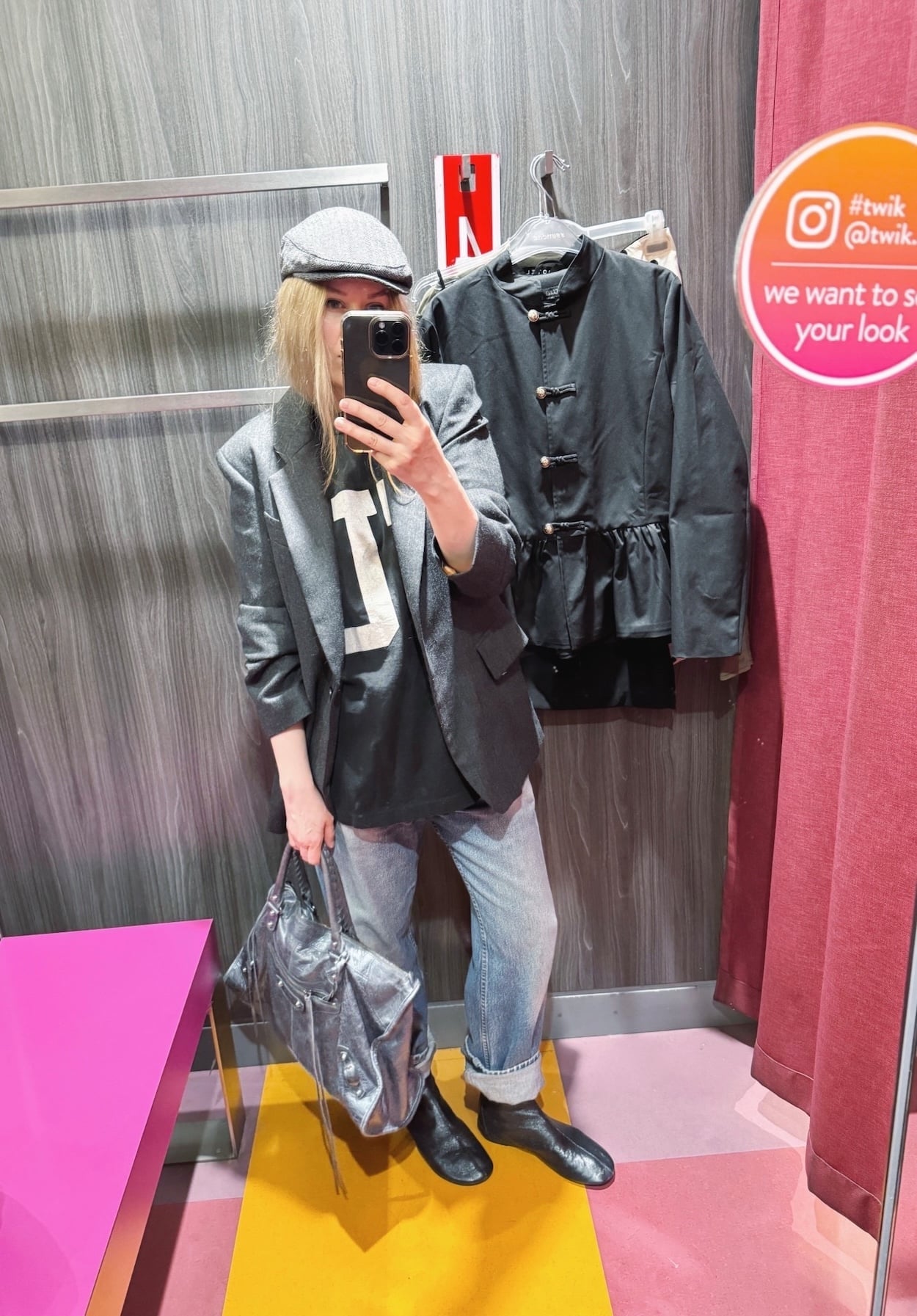 Sara of livelovesara is standing in front of a mirror taking a photo of her outfit. She is wearing a, FU t-shirt under a grey blazer, vintage Levi's. black flat boots, a grey driving cap, and a large grey Balenciaga bag.