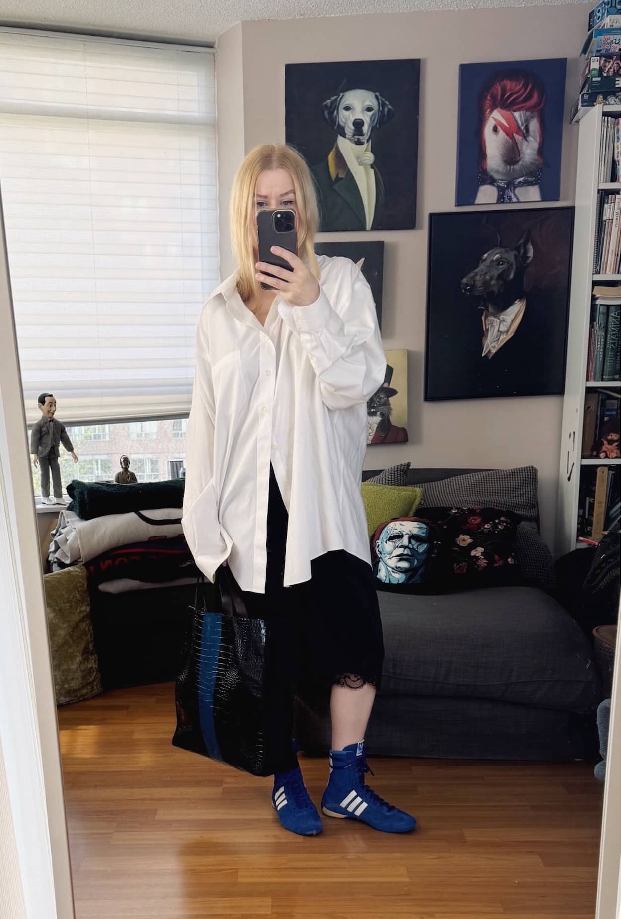 Sara of livelovesara is standing in front of a mirror taking a photo of her outfit. She is wearing an oversized men's white button up, with silk bermuda shorts, blue Adidas wrestling boots, and a tote.