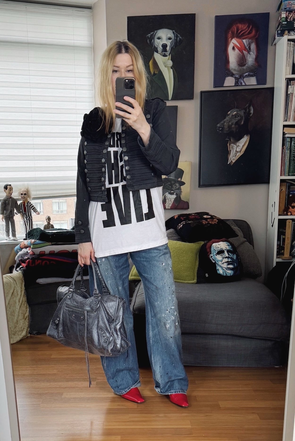 Sara of livelovesara is standing in front of a mirror taking a photo of her outfit. She is wearing a white t-shirt with Saturday Night Live written on it, a grey Napoleon jacket with a latge black flower brooch, jeans with paint splatters, red boots, and a Balenciaga bag in grey.