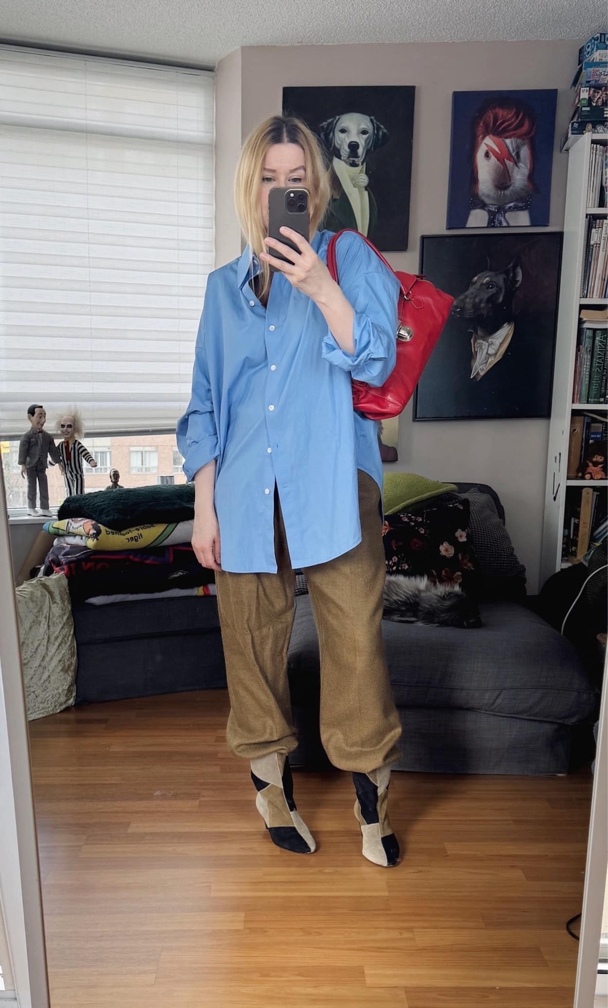 Sara of livelovesara is standing in front of a mirror taking a photo of her outfit. She is wearing a blue button up with camel vintage mens trousers, isabel marant patch boots, and a red y2k Prada bag.