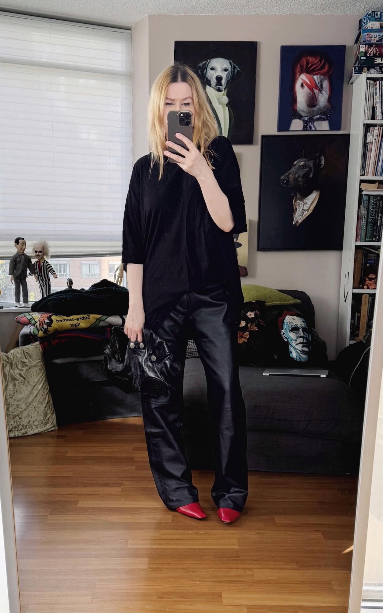 Sara of livelovesara is standing in front of a mirror taking a photo of her outfit. She is wearing a black oversized t-shirt with vintage leather trousers, red boots, and an Alexander McQueen clutch.