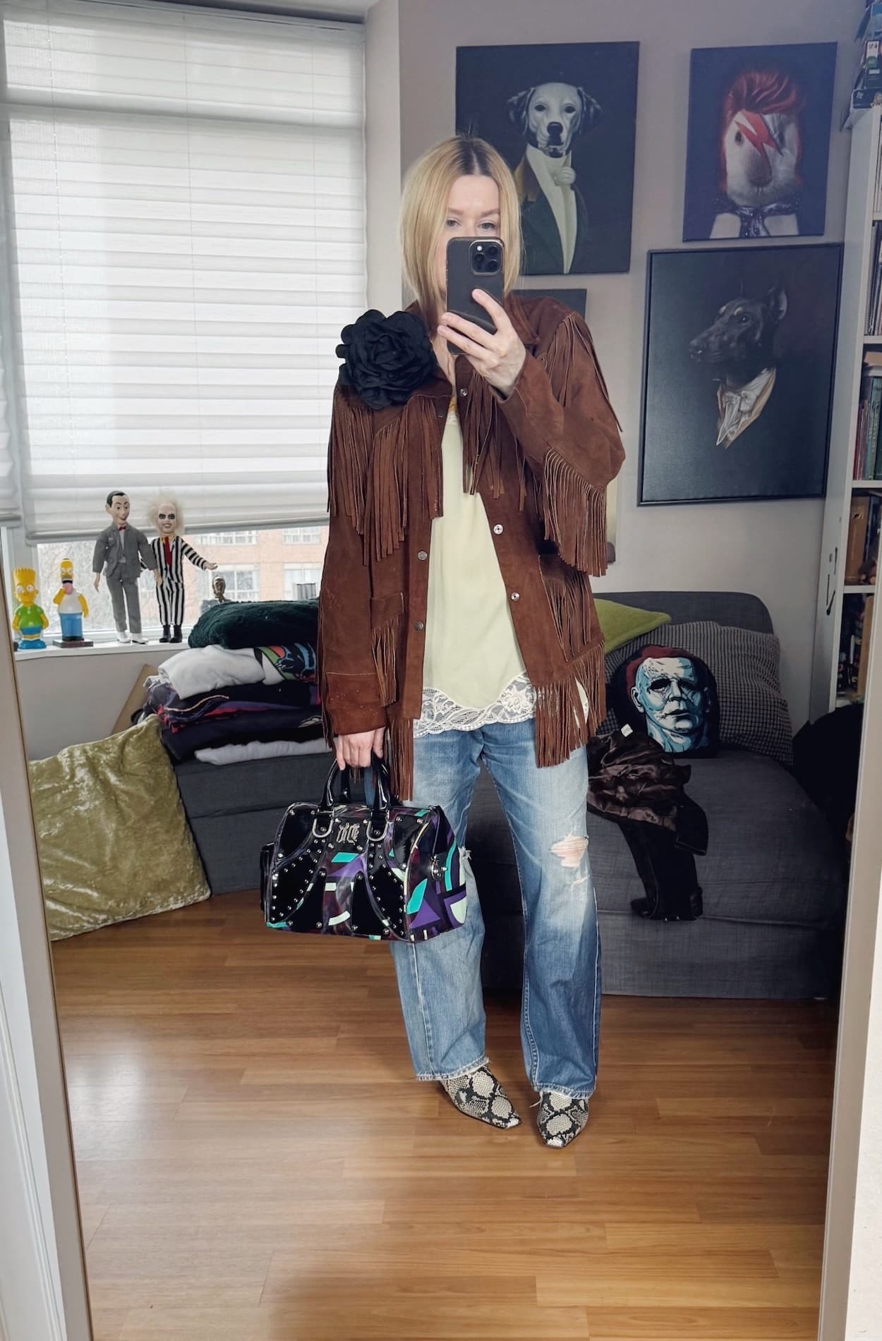 Sara of livelovesara is standing in front of a mirror taking a photo of her outfit. She is wearing a green camisole under a fringe jacket with a large black flower brooch, vintage Levis, snake print boots, and a Y2K Versace bag.