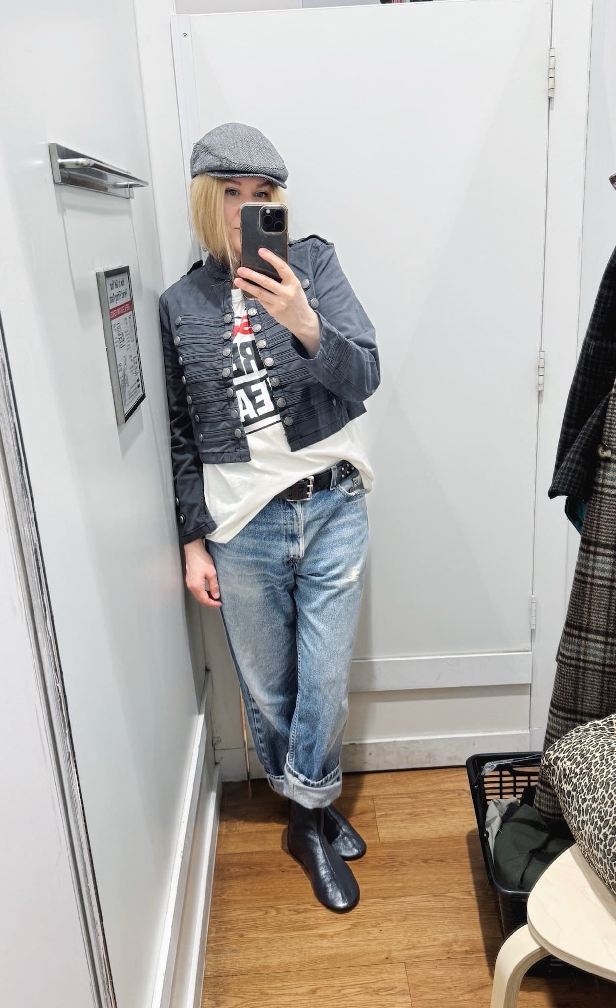 Sara of livelovesara is standing in front of a mirror taking a photo of her outfit. She is wearing a Vision Streetwear tee under a Napoleon jacekt, vintage lives cuffed with a studded belt, flat black boots, and a grey driving cap.