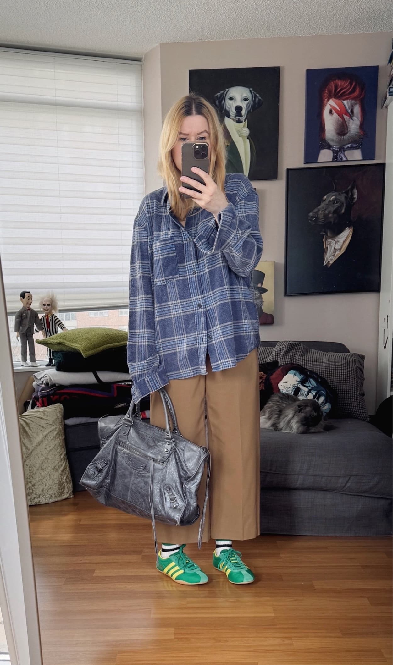 Sara of livelovesara is standing in front of a mirror taking a photo of her outfit. She is wearing a blue plaid shirt with camel cropped trousers, green Adidas, and a large grey Balenciaga work bag.