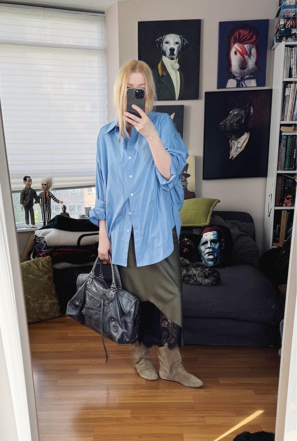 Sara of livelovesara is standing in front of a mirror taking a photo of her outfit. She is wearing an oversized blue button up over and olive green slip dress, tan slouchy boots, and a grey Balenciaga city bag.