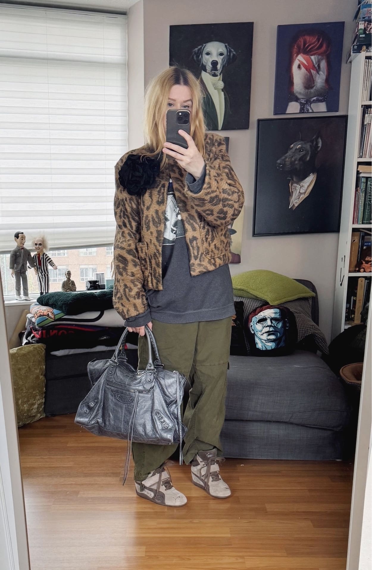 Sara of livelovesara is standing in front of a mirror taking a photo of her outfit. She is wearing a Fugazi sweatshirt under a cropped animal print jacket with a black flower brooch, green cargo pants, puma wedge sneakers, and a large Balenciaga city Work bag.