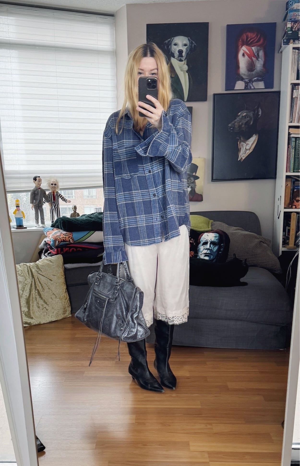 Sara of livelovesara is standing in front of a mirror taking a photo of her outfit. She is wearing an blue plaid shirt with cream satin Bermuda shorts, knee high boots, and a Balenciaga city bag.