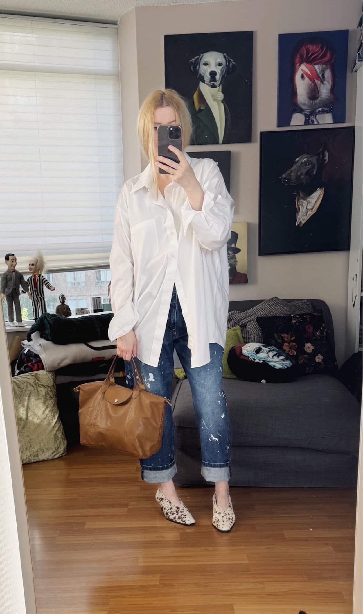 Sara of livelovesara is standing in front of a mirror taking a photo of her outfit. She is wearing a men's oversized white button up with paint splatter jeans, deer print heels, and is carrying a Longchamp Le Plaige bag in brown leather.