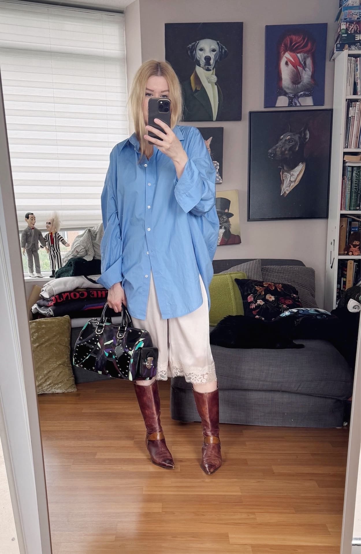 Sara of livelovesara is standing in front of a mirror taking a photo of her outfit. She is wearing an oversized blue button up with cream satin and lace bermuda pants, brown vintage 90s knee high boots, and a Y2K Versace bag.