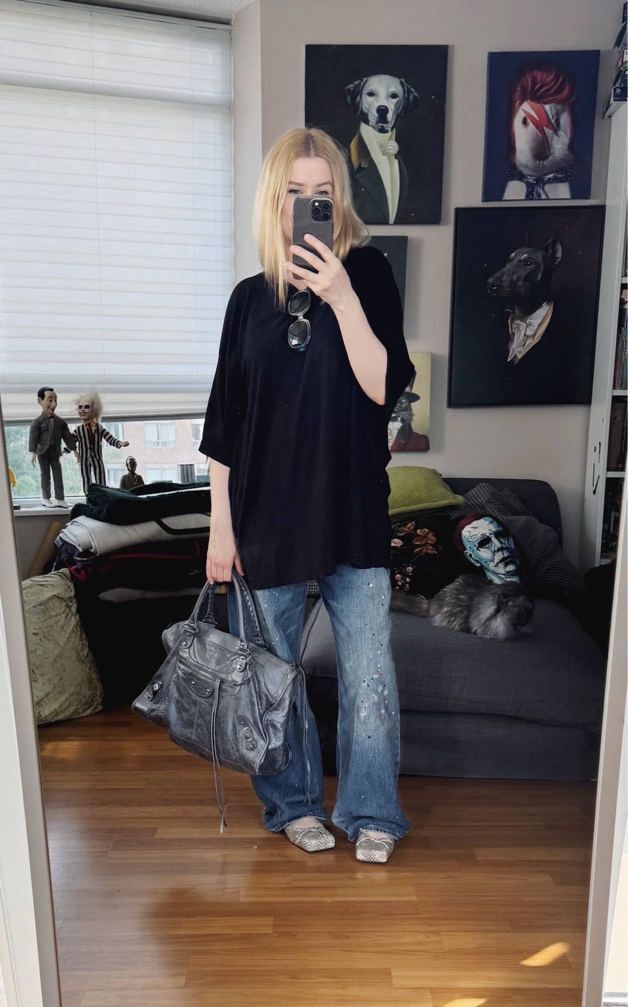 Sara of livelovesara is standing in front of a mirror taking a photo of her outfit. She is wearing an oversized black men's tee with wide leg, paint splattered jeans, square toe, snake print mules, and is has some large vintage sunglasses, and a grey balenciaga city work bag.