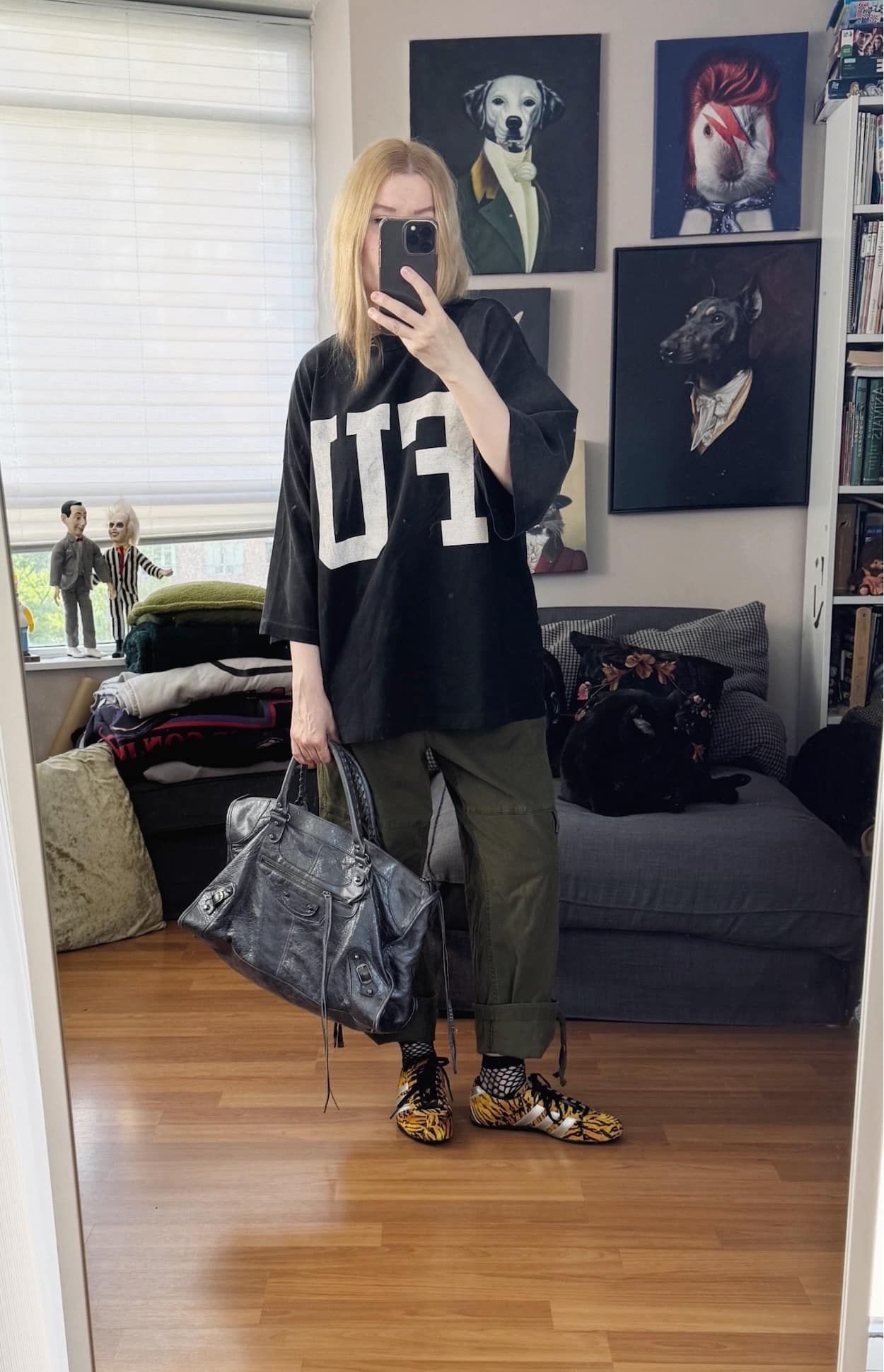 Sara of livelovesara is standing in front of a mirror taking a photo of her outfit. She is wearing an FU oversized t-shirt, cargo pants, tiger print Adidas sneakers, and a grey Balenciaga work bag.