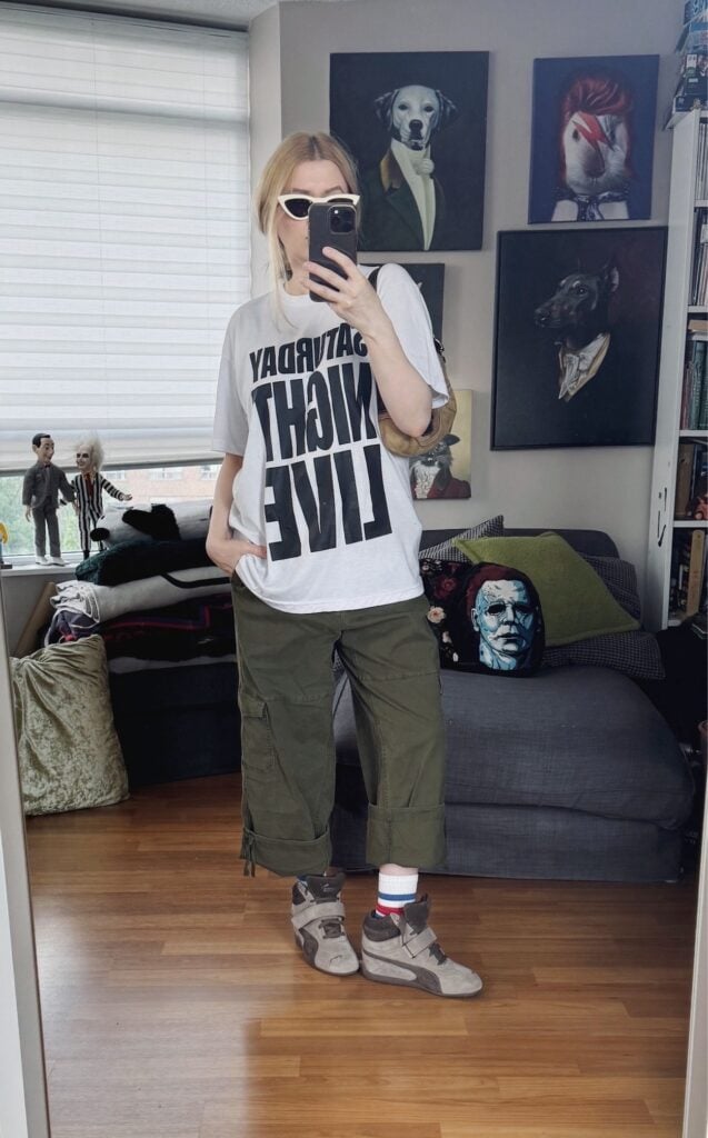 Sara of livelovesara is standing in front of a mirror taking a photo of her outfit. She is wearing a white Saturday Night Live T-shirt with green cargos that are cuffed to culotte length, cream cat eye sunglasses, Puma wedge sneakers, and is carrying a Y2K coach soho bag.