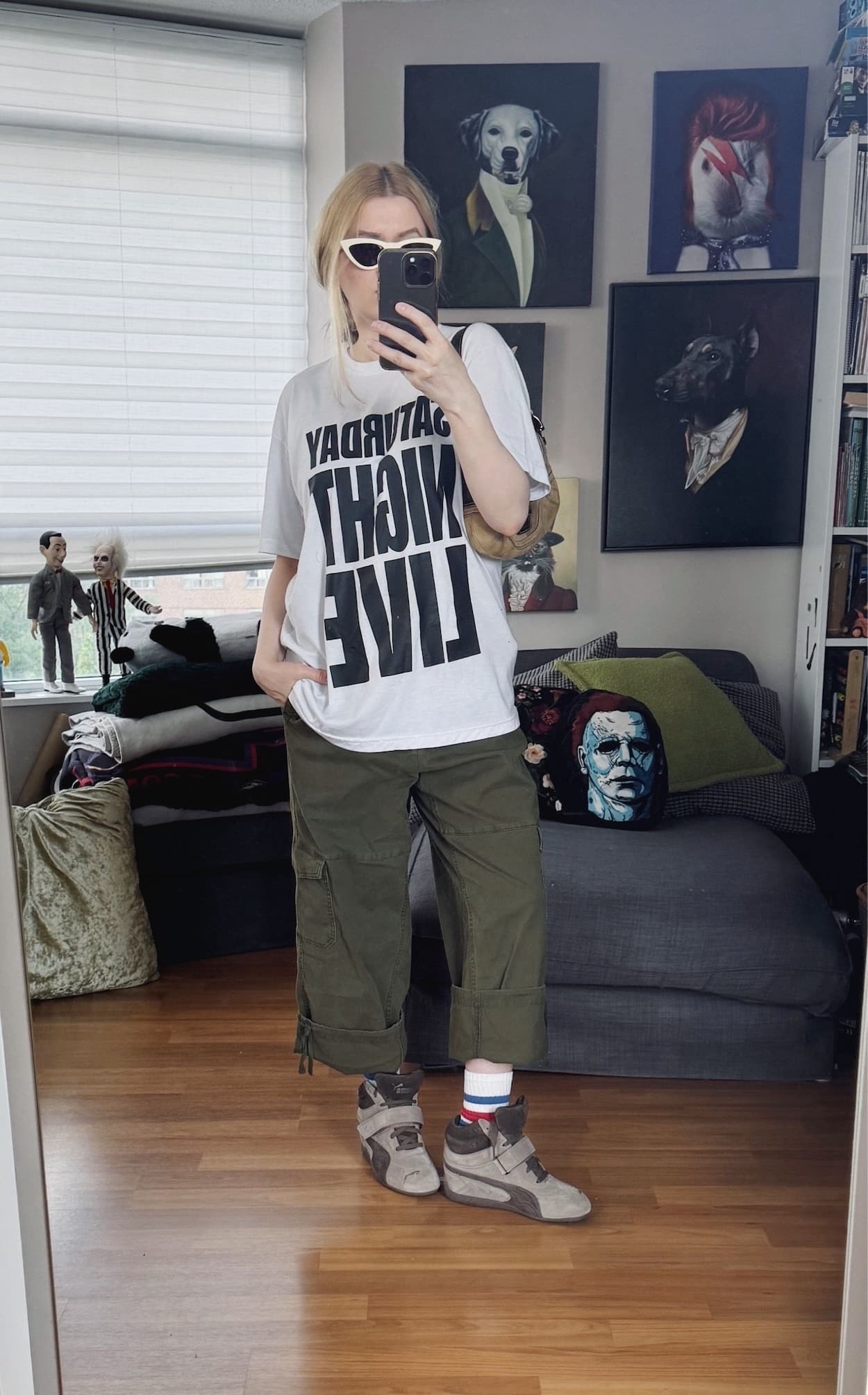 Sara of livelovesara is standing in front of a mirror taking a photo of her outfit. She is wearing a white Saturday Night Live T-shirt with green cargos that are cuffed to culotte length, cream cat eye sunglasses, Puma wedge sneakers, and is carrying a Y2K coach soho bag.