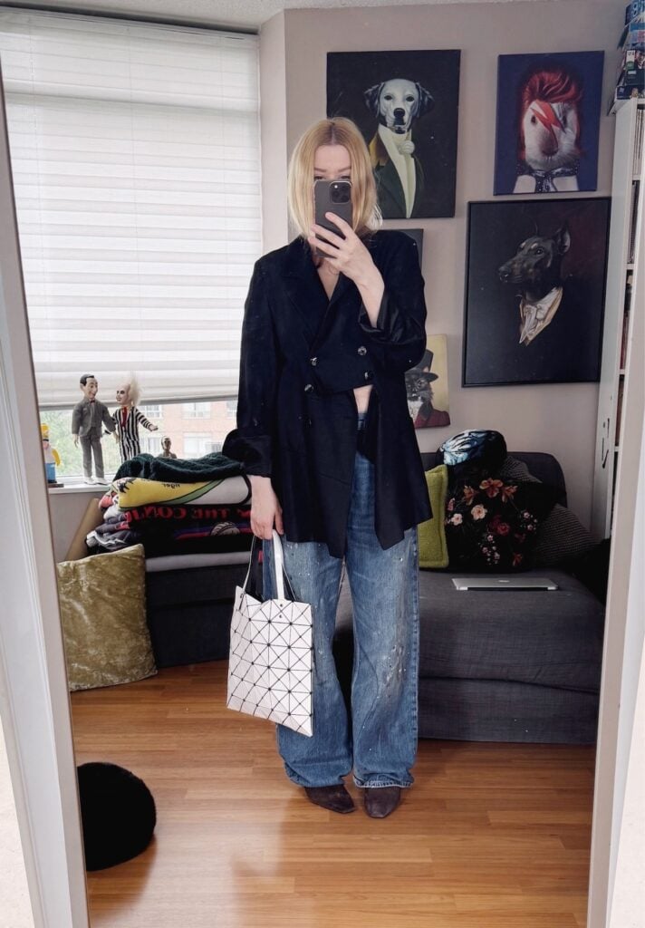 Sara of livelovesara is standing in front of a mirror taking a photo of her outfit. She is wearing a black vintage deconstructed linen blazer with paint splatter jeans, brown suede mules, and a white Issey Miyake Bao Bao bag.