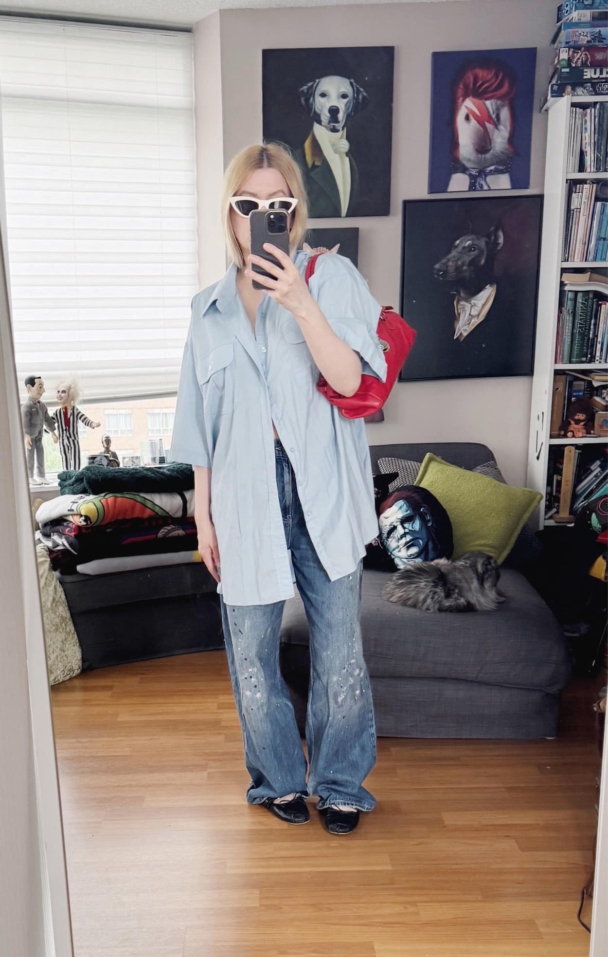 Sara of livelovesara is standing in front of a mirror taking a photo of her outfit. She is wearing an oversized light blue, short sleeve button up, over wide leg, paint splatter jeans, black Miu Miu ballet flats, cream cat eye sunglasses by Celine, and is carrying a red vintage Prada bag.