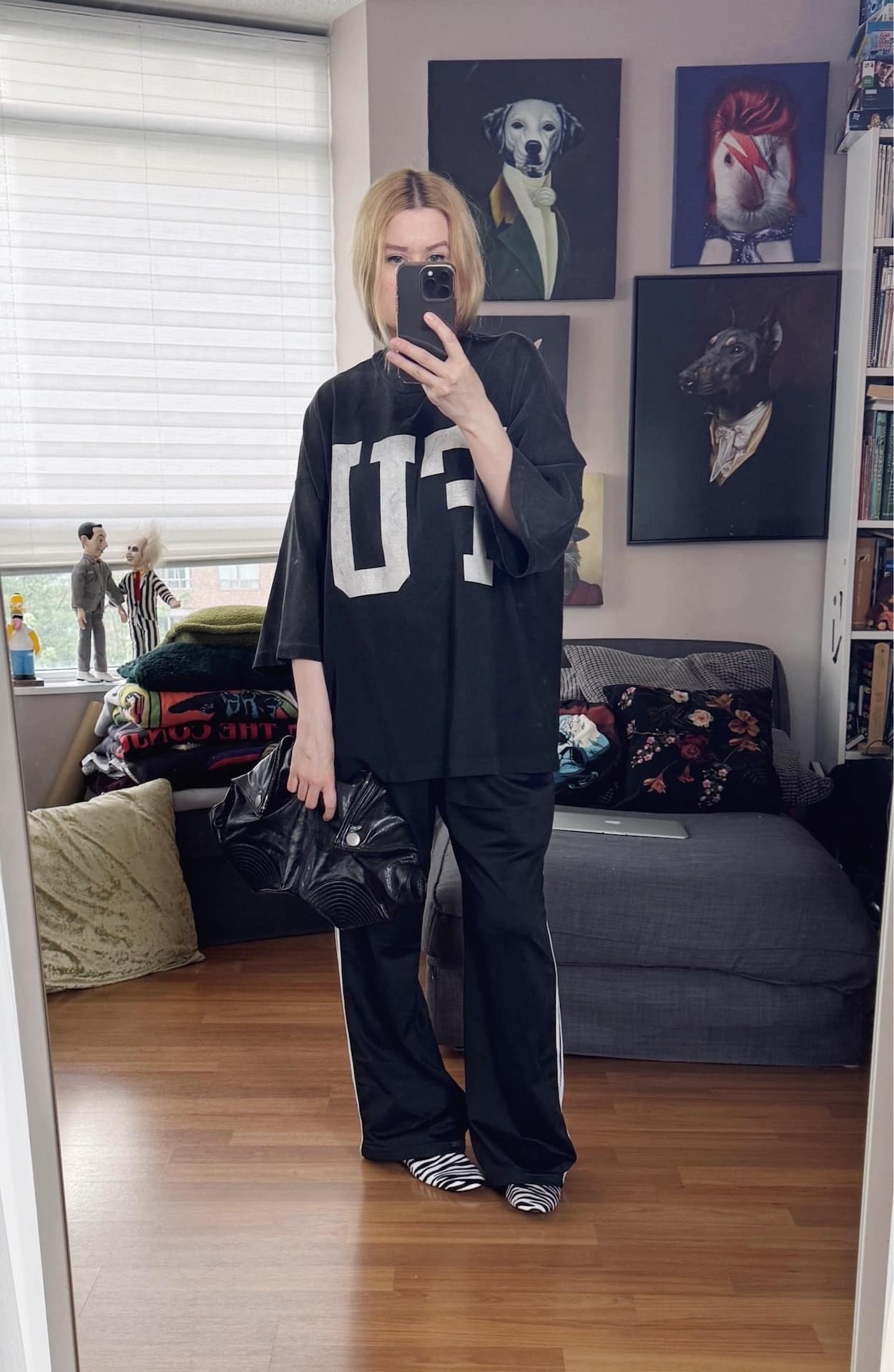 Sara of livelovesara is standing in front of a mirror taking a photo of her outfit. She is wearing a black t-shirt with a large FU on the front, paired with black Adidas track pants, zebra print loafers, and a black Alexander McQueen de manta clutch.