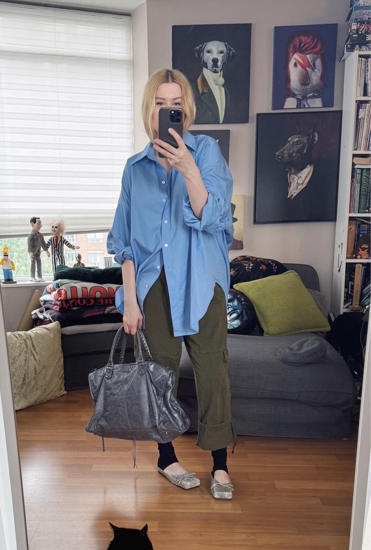 Sara of livelovesara is standing in front of a mirror taking a photo of her outfit. She is wearing an oversized blue button up with cuffed green cargo pants, half socks, snake print mules, and is carrying a large, grey balenciaga city work bag.