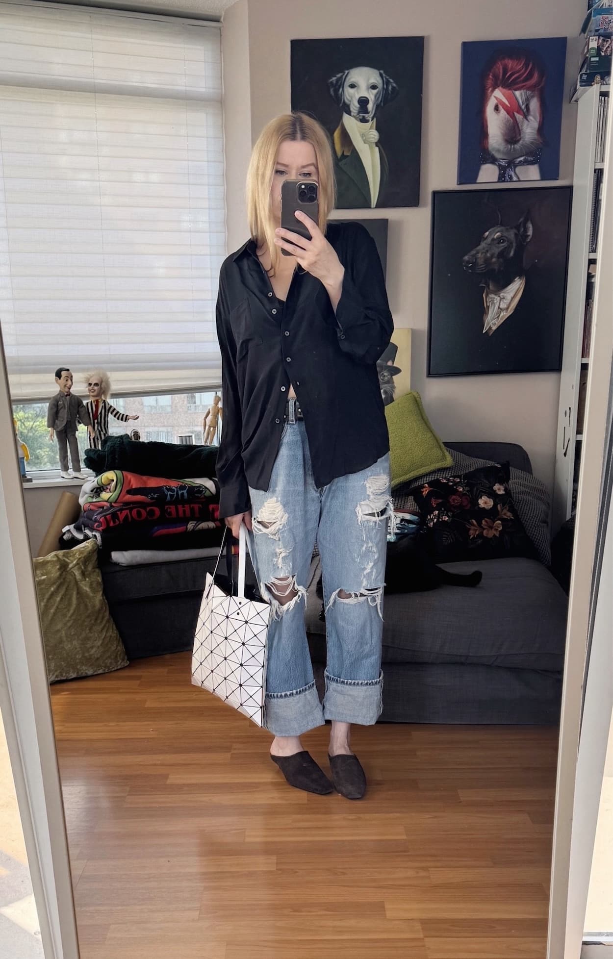 Sara of livelovesara is standing in front of a mirror taking a photo of her outfit. She is wearing a black silk shirt, distressed vintage levis, brown swead wedge mules, and is carrying a white Issey Miyake Bao Bao bag.