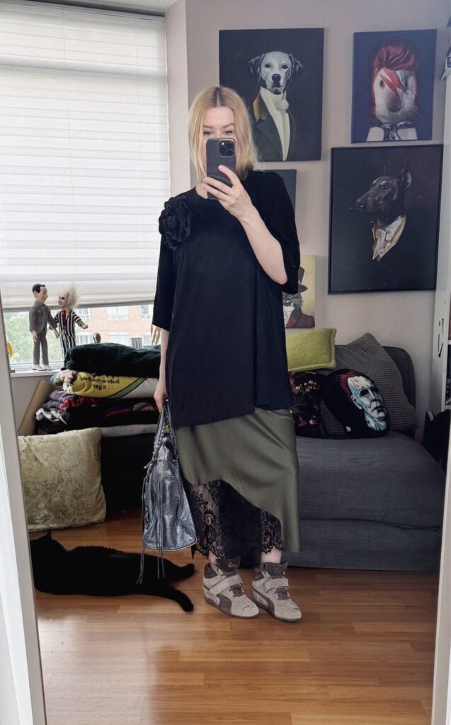 Sara of livelovesara is standing in front of a mirror taking a photo of her outfit. She is wearing an olive green slip dress with an oversized black t-shirt over it. brown Puma wedge sneakers, a black flower brooch, and is carrying a large, grey Balenciaga work bag.