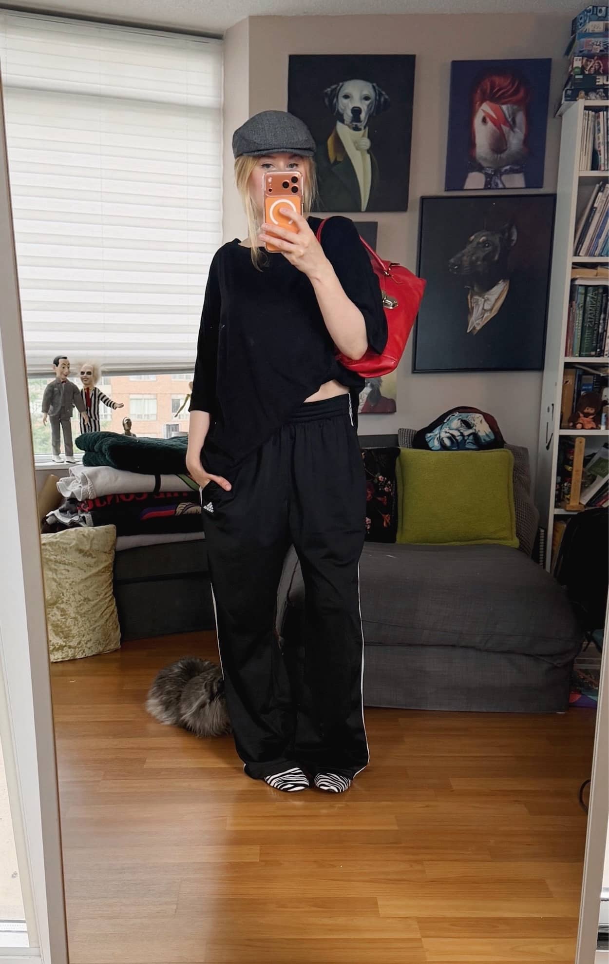 Sara of livelovesara is standing in front of a mirror taking a photo of her outfit. She is wearing an oversized black t-shirt, Adidas track pants, zebra print loafers, a red Prada bag, and a grey herringbone flat cap.