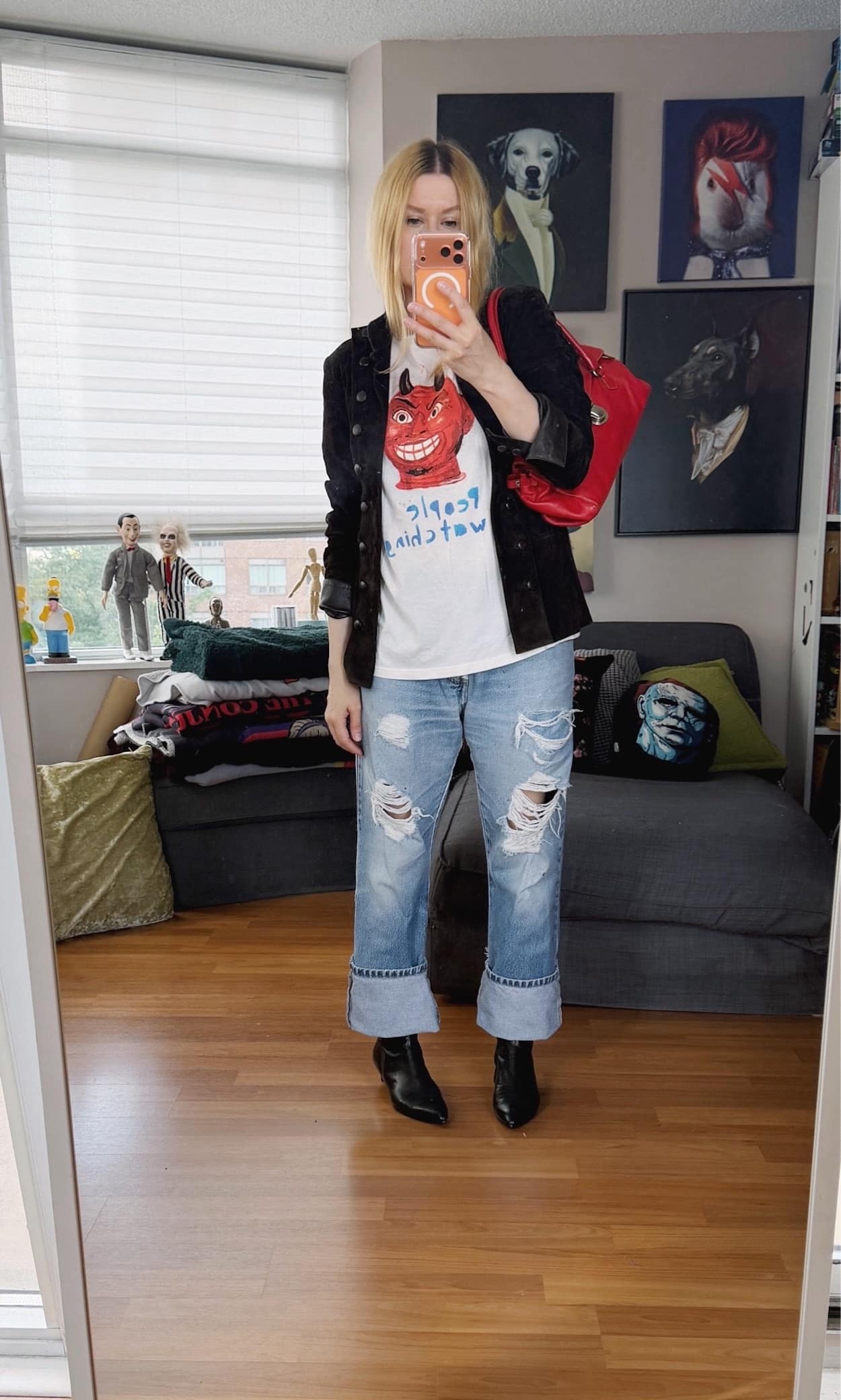 Sara of livelovesara is standing in front of a mirror taking a photo of her outfit. She is wearing a white tee that says People Watching under a cartoon devil, vintage Levis, a brown suede Napoleon jacket, black boots, and is carrying a red y2k Prada bag.