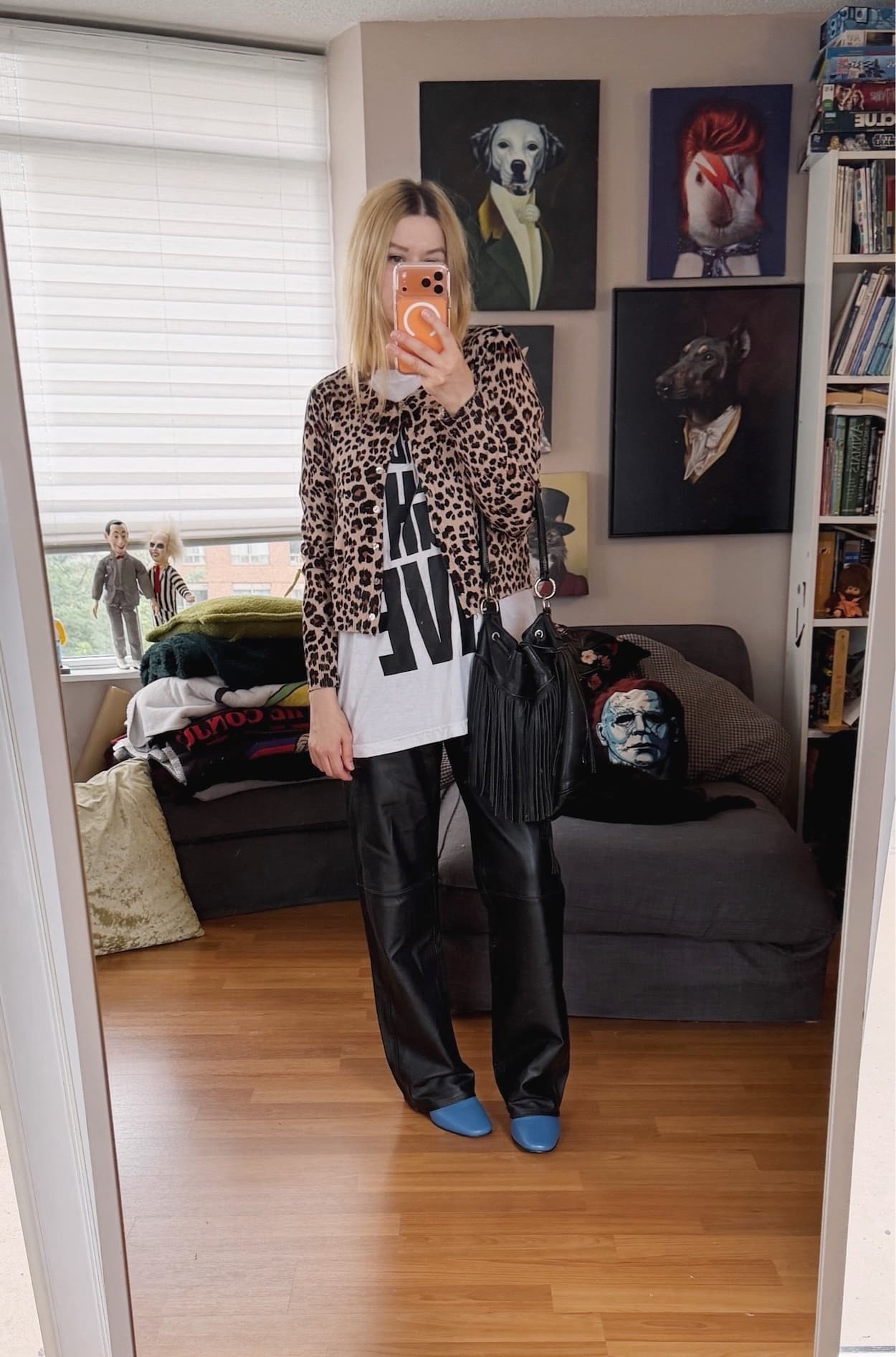 Sara of livelovesara is standing in front of a mirror taking a photo of her outfit. She is wearing a white Saturday Night Live tee under an animal print cardigan, vintage leather trousers, blue boots, and a fringe bucket bag.