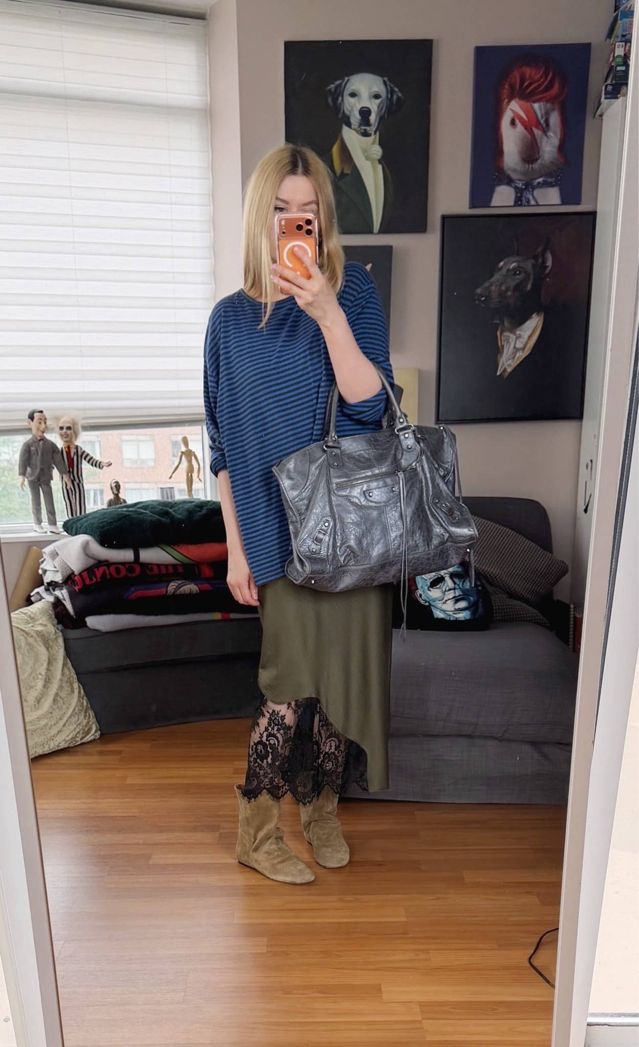 Sara of livelovesara is standing in front of a mirror taking a photo of her outfit. She is wearing an olive green slip dress under a blue striped shirt, brown slouchy boots, and a large Balenciaga work bag.