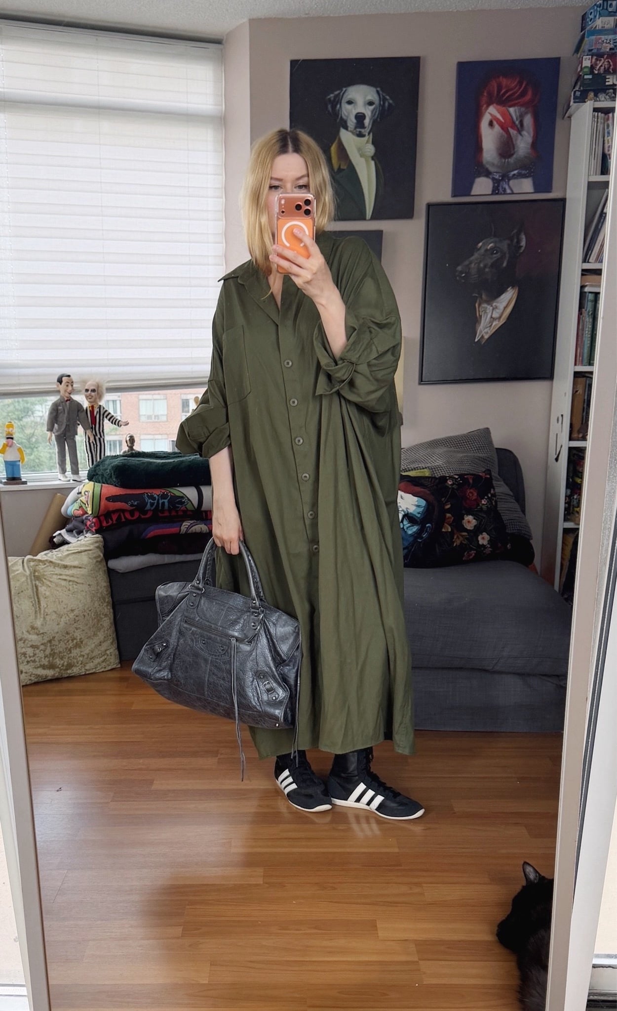 Sara of livelovesara is standing in front of a mirror taking a photo of her outfit. She is wearing a very oversized green shirt dress, black Adidas boxer sneakers, and is carrying a large Balenciaga city work bag.