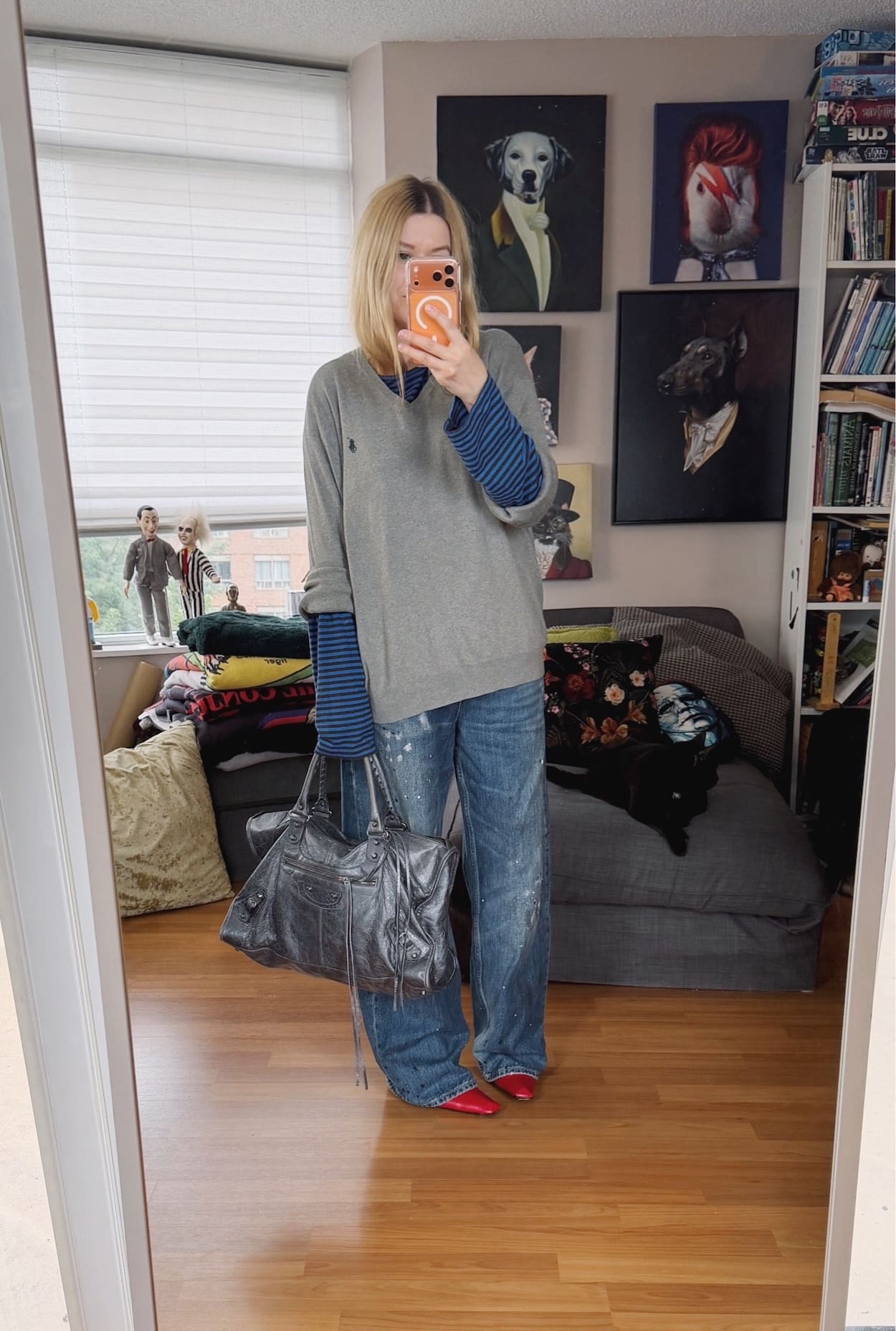 Sara of livelovesara is standing in front of a mirror taking a photo of her outfit. She is wearing a blue striped long sleeve under a grey Ralph Lauren sweater, oversized paint splatter jeans, red vintage boots, and is carrying a large grey Balenciaga work bag.