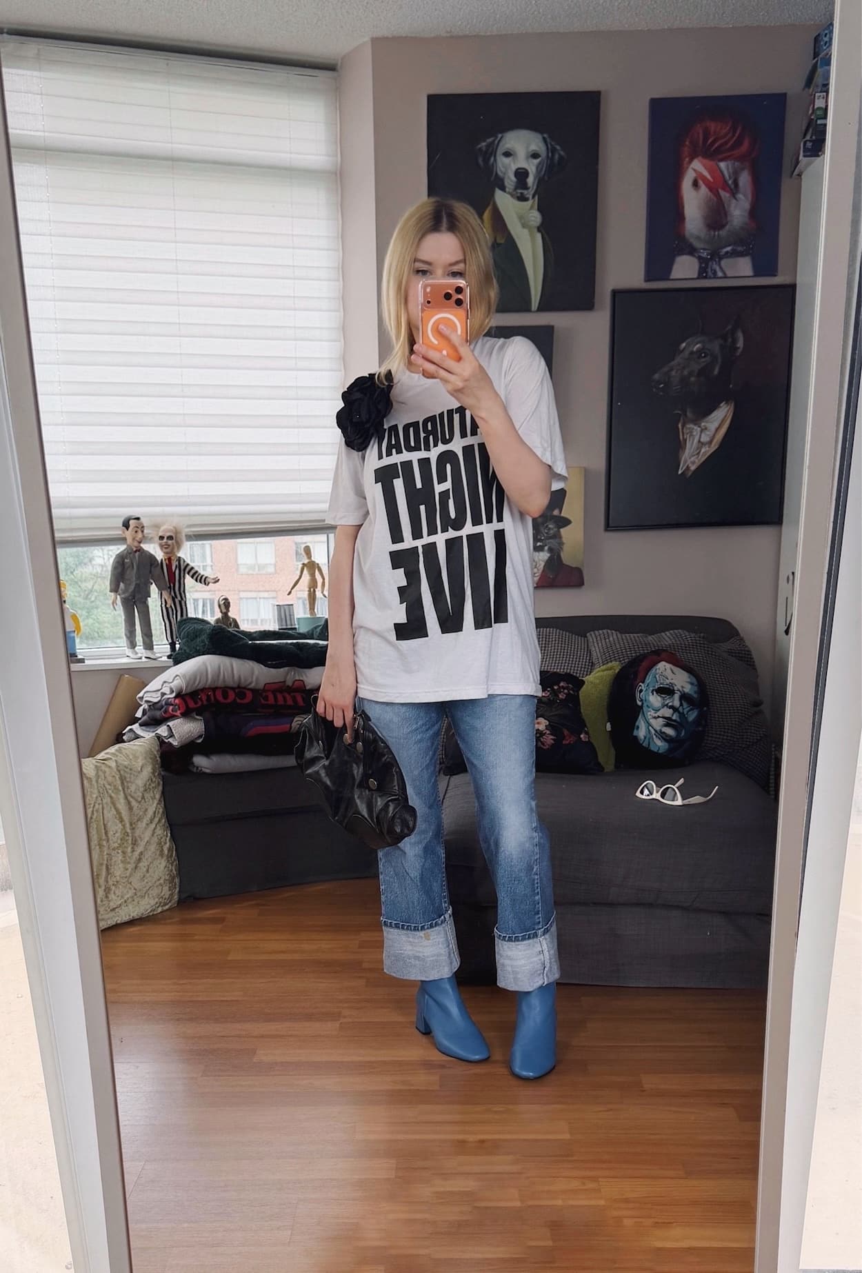 Sara of livelovesara is standing in front of a mirror taking a photo of her outfit. She is wearing an oversized white Saturday Night Live tee with a black flower brooch, vintage Levis cuffed over blue boots, and an Alexander McQueen clutch.