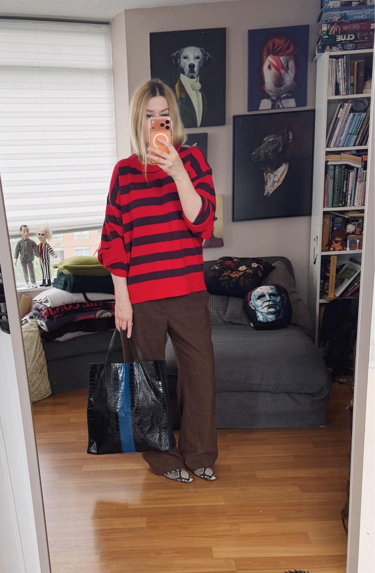Sara of livelovesara is standing in front of a mirror taking a photo of her outfit. She is wearing a red striped sweater with vintage brown trousers, snake print boots, and is carrying a black tote with a blue central stripe.