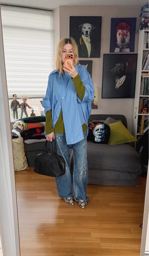 Sara of livelovesara is standing in front of a mirror taking a photo of her outfit. She is wearing a green striped long sleeve under a sky blue button up. Paint splstter wide leg jeans, with snake print flat boots, and is carrying a black leather YSL easy bag.