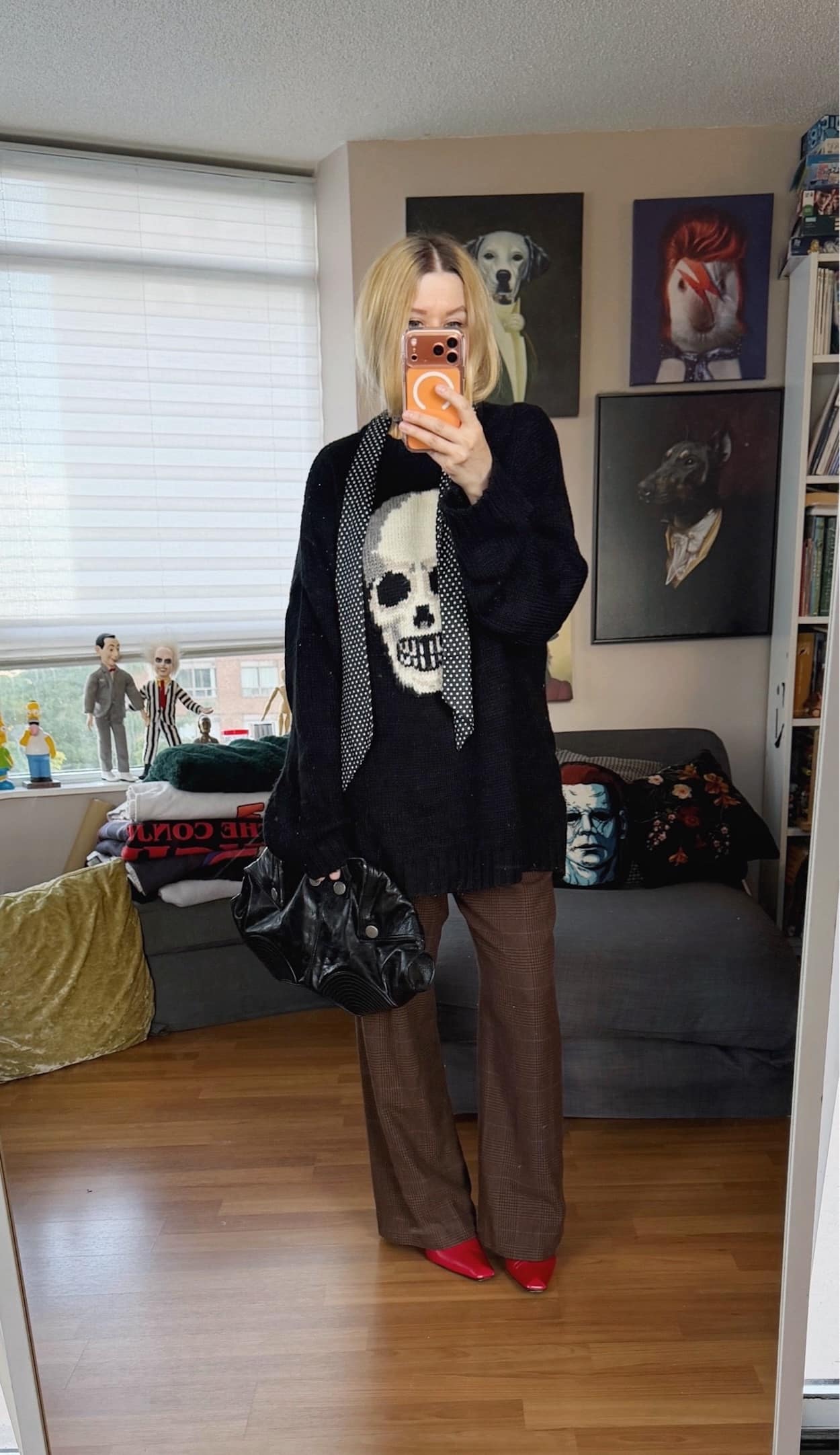 Sara of livelovesara is standing in front of a mirror taking a photo of her outfit. She is wearing a black sweater with a skull, a polka dot skinny scarf, brown plaid vintage trousers, red vintage boots, and an ALexander McQueen clutch.
