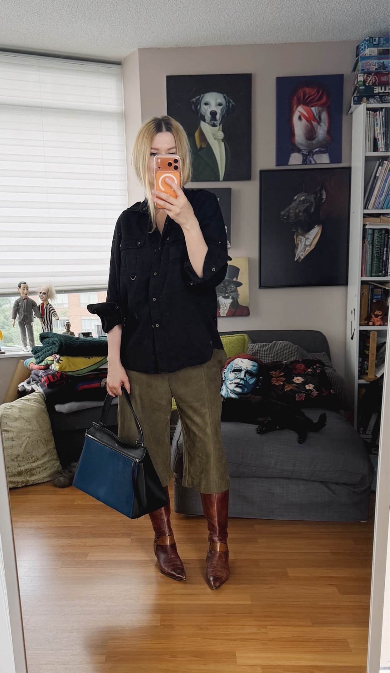 Sara of livelovesara is standing in front of a mirror taking a photo of her outfit. She is wearing a black Ralph Lauren utility shirt with green fake suede Bermuda shorts, tall brown vintage boots, and is carrying a blue celine edge bag.