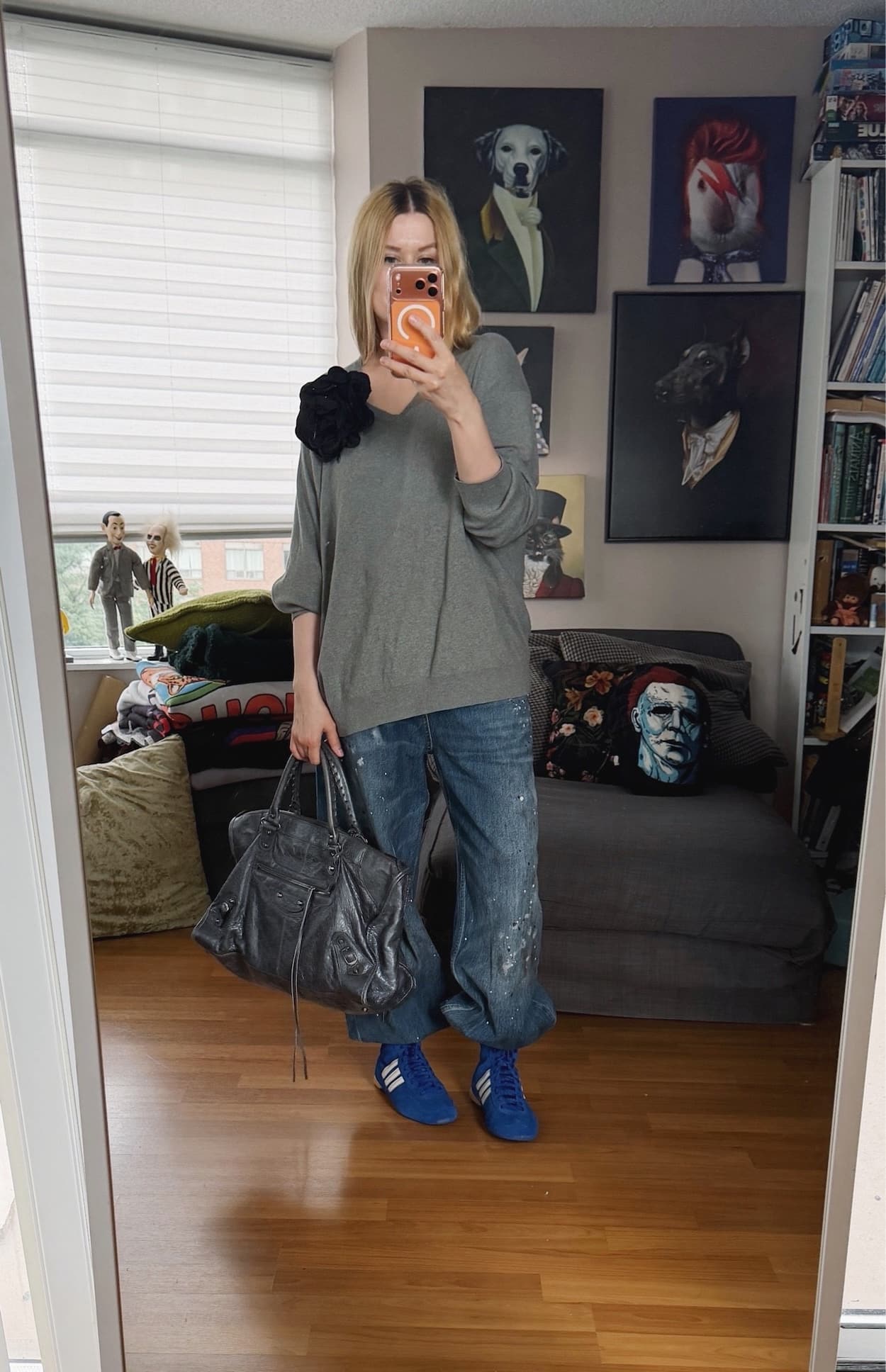 Sara of livelovesara is standing in front of a mirror taking a photo of her outfit. She is wearing a grey vintage Ralph Lauren sweater with a black flower brooch, paint splattered jeans tucked into blue Adidas wrestling boots, and a grey Balenciaga work bag.