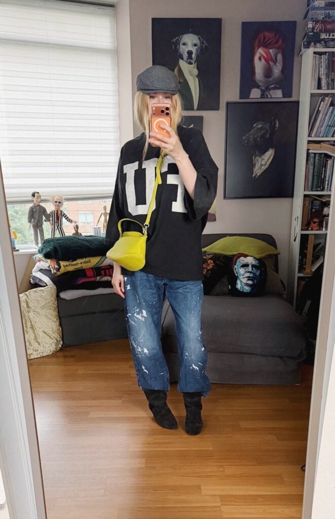 Sara of livelovesara is standing in front of a mirror taking a photo of her outfit. She is wearing a Fear of God FU t-shirt with paint splatter jeans tucked into Isabel Marant slouchy boots, a grey flat cap, and a neon yellow crossbody.