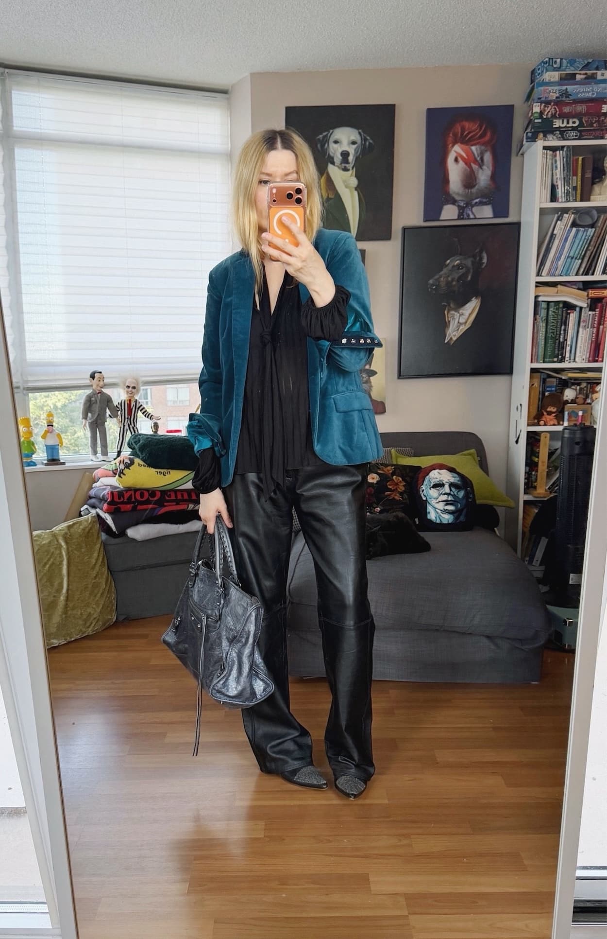 Sara of livelovesara is standing in front of a mirror taking a photo of her outfit. She is wearing a black tie neck shirt under a blue velvet blazer, vintage leather trousers, black boots, and is carrying a grey Balenciaga bag.