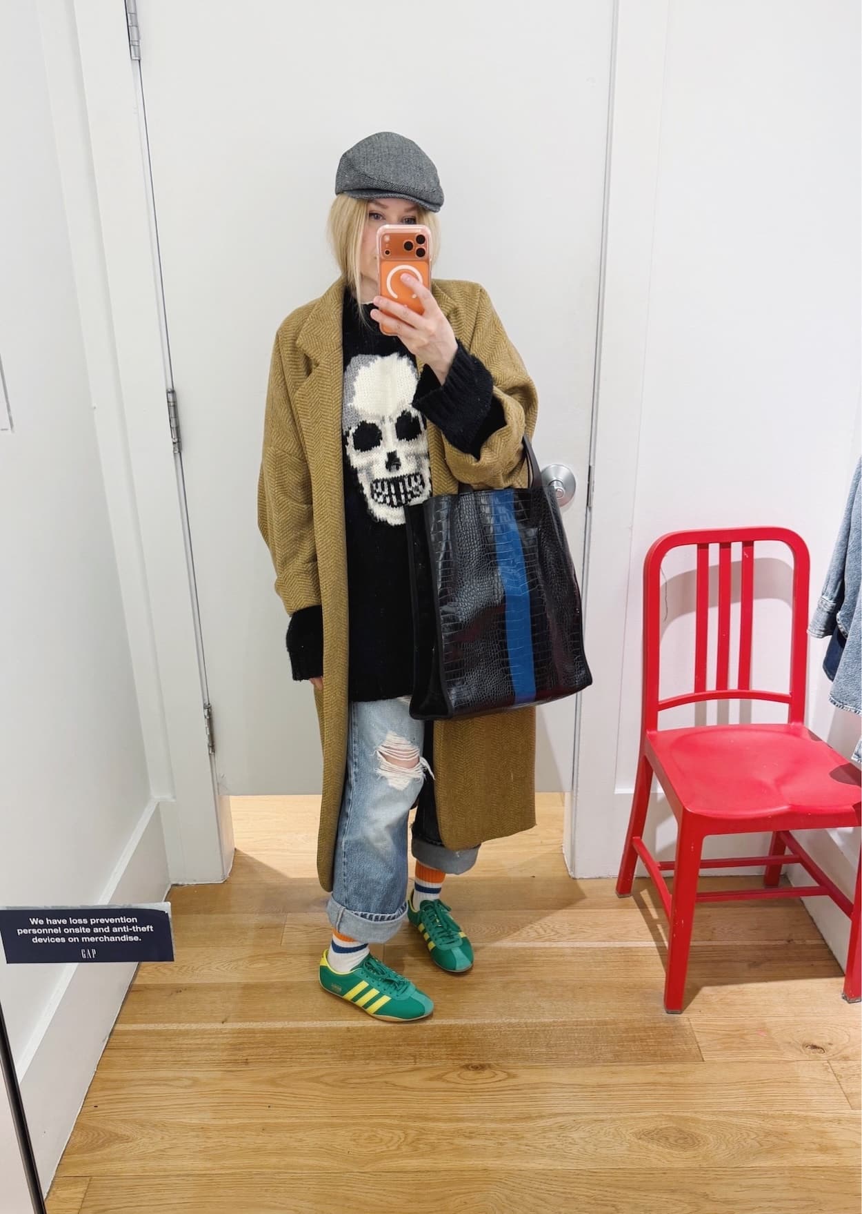 Sara of livelovesara is standing in front of a mirror taking a photo of her outfit. She is wearing a black skull sweater under a Horses Atelier coat, vintage Levis, green Adidas a grey herringbone cap, and a black tote with a blue stripe.