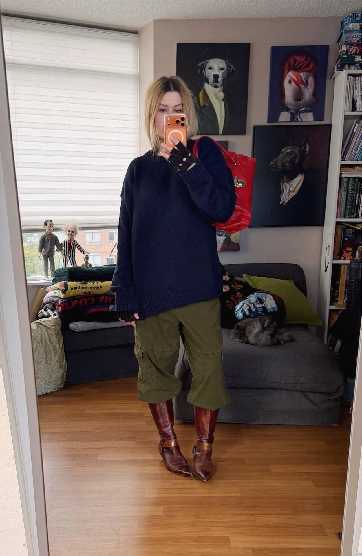 Sara of livelovesara is standing in front of a mirror taking a photo of her outfit. She is wearing a navy blue oversized, cotton Gap sweater with green cargo pants tucked into 90s brown boots, suede fingereless gloves, and a red prada bag.