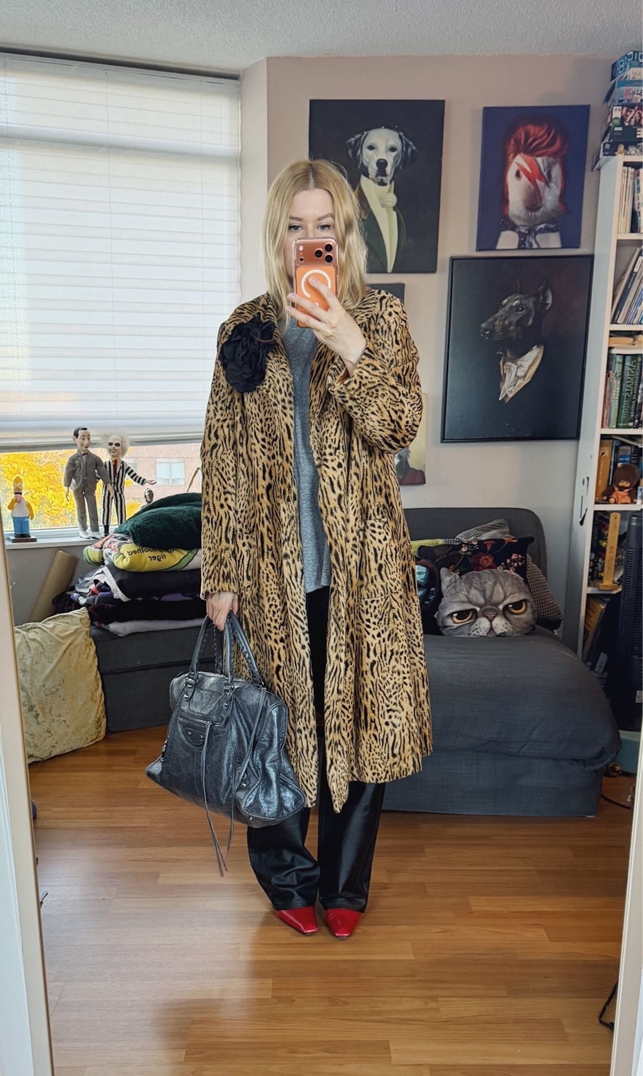 Sara of livelovesara is standing in front of a mirror taking a photo of her outfit. She is wearing a grey t-shirt with vintage leather pants, a vintage animal print coat with a black flower brooch, red heeled boots, and is carrying a large grey Balenciaga work bag.