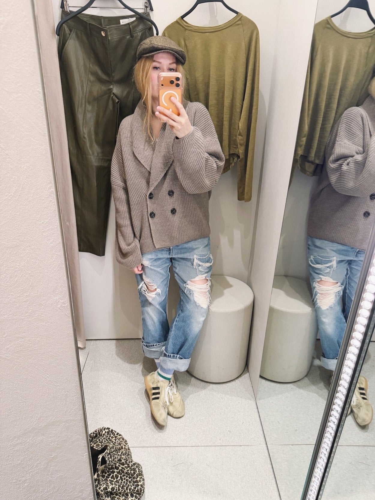 Sara of livelovesara is standing in front of a mirror taking a photo of her outfit. She is wearing a grandpa cardigan with distressed vintage levis, a brown flat cap, and SFTM x Adidas tan sneakers with an animal print tote bag.