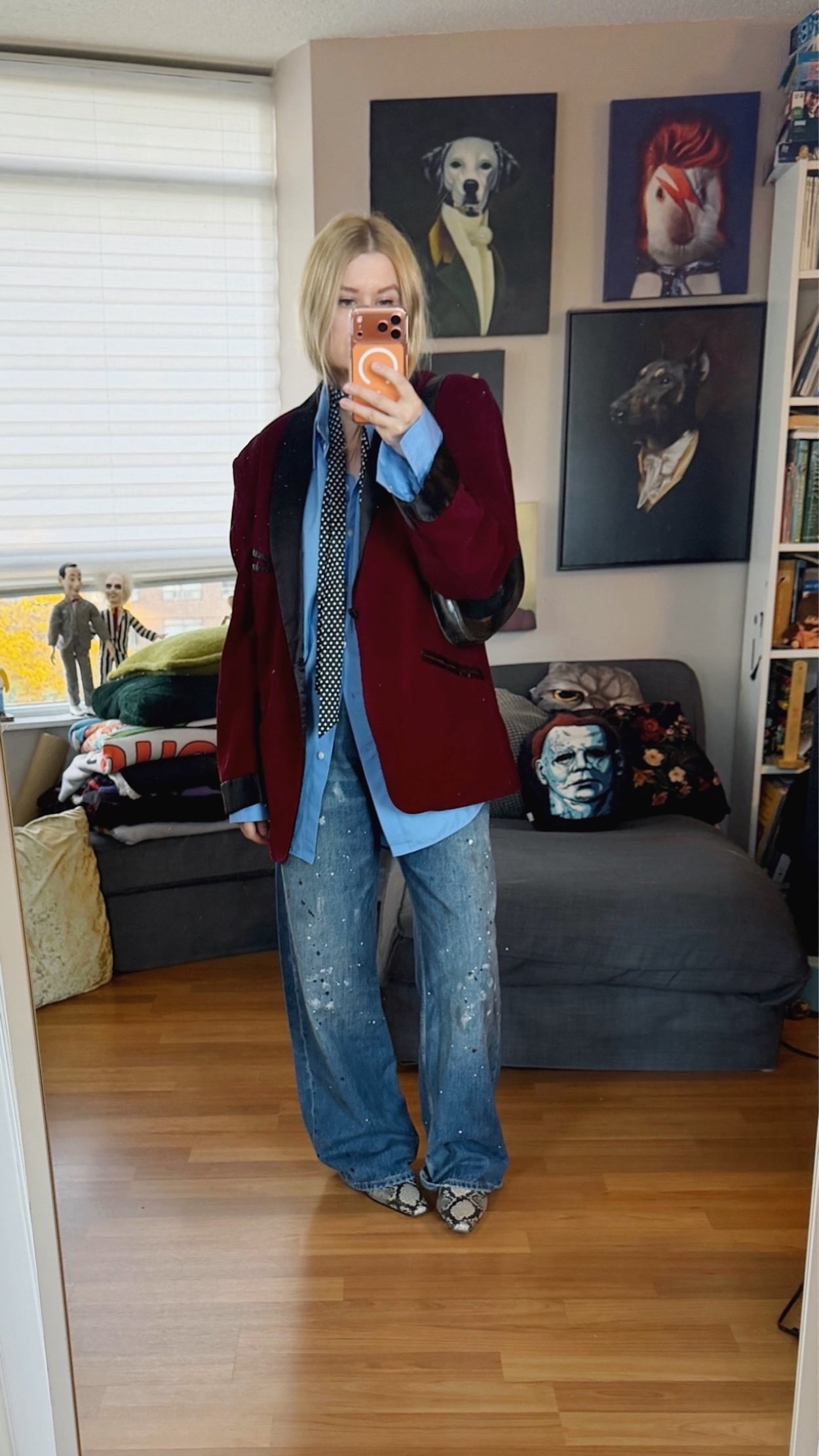 Sara of livelovesara is standing in front of a mirror taking a photo of her outfit. She is wearing a blue button up under a men's burgundy velvet vintage smoking jacket, a polka dot skinny scarf, paint splatter jeans, snake print boots, and is carrying a vintage Gucci jackie bag.